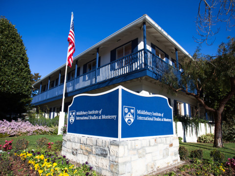 A doublesided blue and white sign reading "Middlebury Institute of International Studies at Monterey" sits below a white building with blue trim and blue fencing along the upstairs walkway.