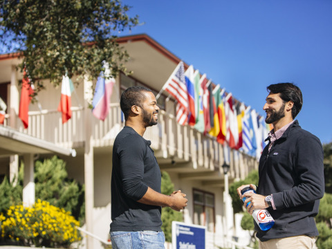 Two men wearing black longsleeve shirts smile as they converse outside a two-story building with various countries' flags hanging over the balcony.
