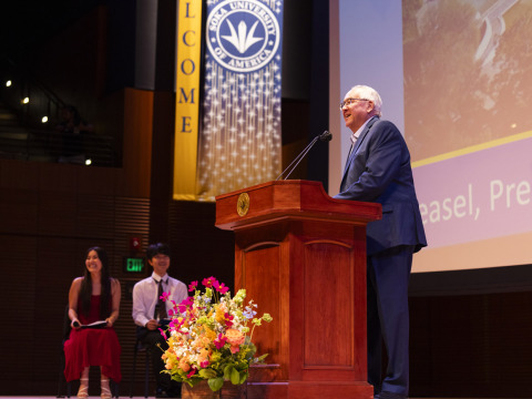 President Feasel wears a suit and speaks into the microphone from behind a wooden lectern. Two students are seated off to the left.