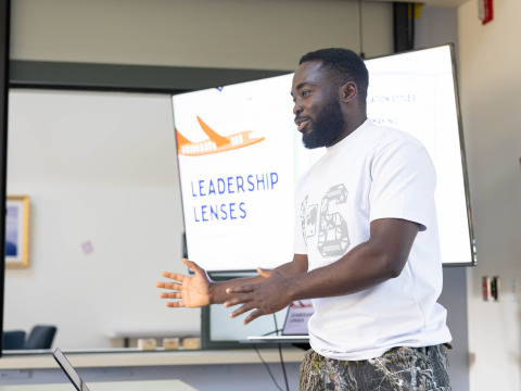 A man wearing a white t-shirt and camo pants gestures with his hands as he stands in front of a projector screen that reads, "Leadership Lenses."