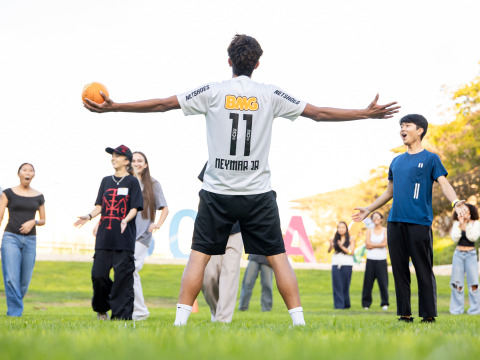 A student with short brown hair wearing a white Neymar jersey stands with his back to the camera and arms outstretched, holding a dodgeball in one hand as other dodgeball players look on.
