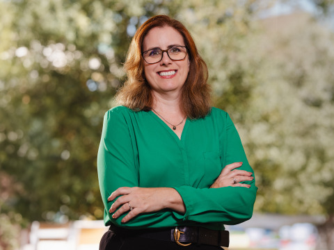 A woman with reddish-brown, shoulder-length hair is wearing a light green blouse, black pants, and black-rimmed glasses as she stands with her arms crossed and smiles. 
