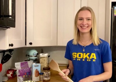 Blond woman wearing blue and yellow Soka T-shirt smiling in kitchen holding wooden spoon