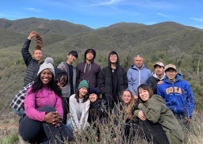 Twelve students and their professor pose together in front of green rolling hills.