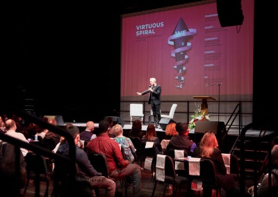 Simon Mainwaring holds a microphone and stands on stage in the Black Box Theatre on Soka's campus where he speaks to an audience about Rewiring Capitalism. There is a screen behind projecting some of his presentation points.