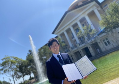 Mark Okuda, winner of the Frederick Douglass Global Fellowship, poses with his Ikeda Scholarship in front of Peace Lake and Founders Hall