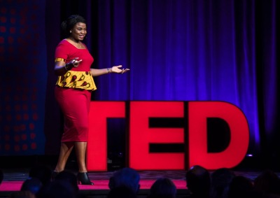 Professor Chika Esiobu stands in front of big red letters spelling out TED as she speaks on stage during her TED Talk