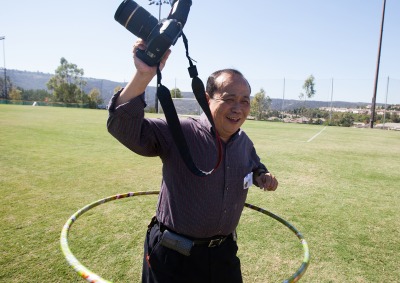 Mitsu Kimura smiles and holds his camera up as he is hula hooping