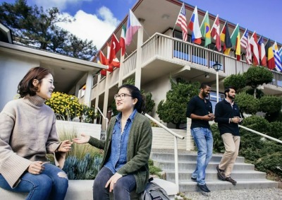 Two female students talk while sitting on a bench beneath a building displaying flags from various countries. Two men are walking down the steps to the side of the women talking.