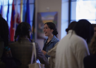 A brunette woman wearing a jean jacket smiles as she speaks with other students in front of a variety of international flags.