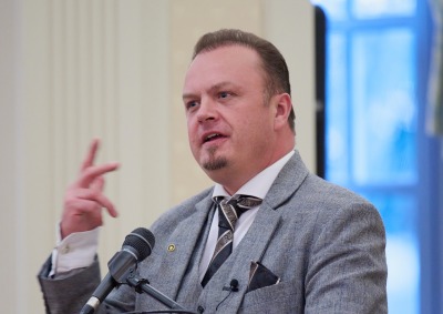 Alexander Harang gestures with his hand as he speaks at a podium with a microphone. He is wearing a grey three piece suit with a black tie.