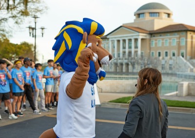 Leo the Lion speaks with Erica Baldaray in front of Founders Hall and Peace Lake before the start of the Heart of a Lion 5k.