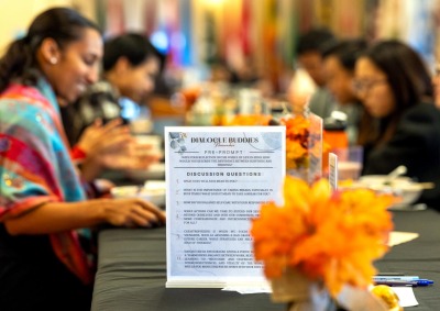 A "Dialogue Buddies" event features reflective questions on wellness and self-care, with participants gathered around a table decorated with orange flowers.