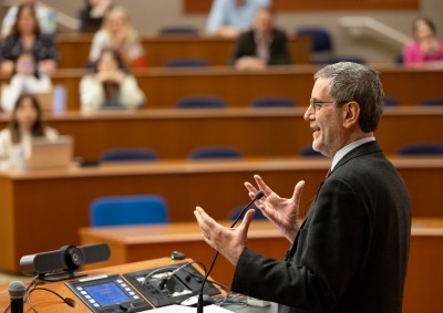 Andrea Bartoli, wearing a suit and glasses, stands at a lectern and gestures with his hands while speaking to an auditorium of people.