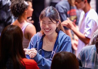 A girl wearing a blue zip-up sweater smiles at another student as they participate in an icebreaker activity.