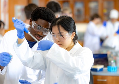 A male and female student in lab coats and safety goggles work together with a micropipette in a science lab.