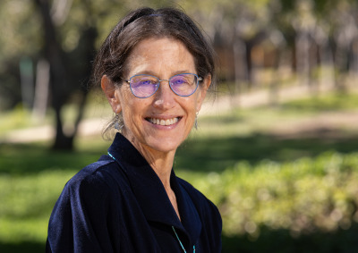 A dark-haired woman wearing a navy blue collared shirt and blue-rimmed glasses smiles at the camera outside in a grassy area.