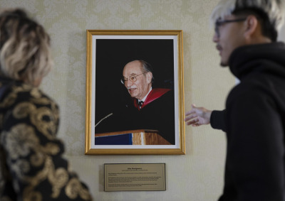 A male student with bleach blonde and black hair gestures towards a framed photo of a man speaking at a lectern.