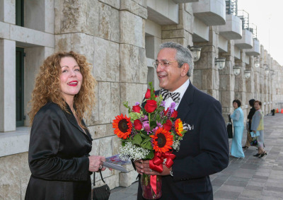 Linda and Luis Neieves are wearing black formal attire, smiling, and Luis is holding a bouquet of red and purple flowers as they stand outside of Founders Hall.