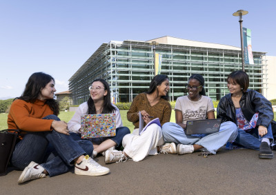 Five female students sit on the sidewalk in front of the Performing Arts Center, some with laptops in their laps, as they smile and laugh with eachother.