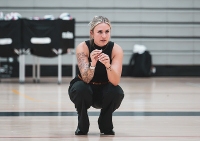 A blonde woman with her hair up, wearing a black tanktop and pants squats down on a gym floor with volleyballs in the distance.