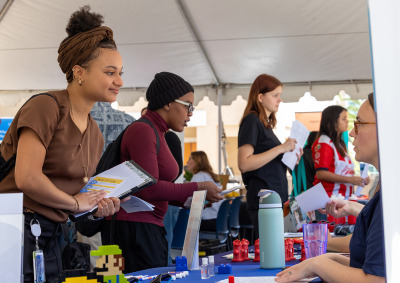A young woman with a brown headwrap and gold jewelry smiles while leaning over a table at an outdoor career fair, holding a clipboard and documents. Other students and recruiters are visible under a large white tent in the background.