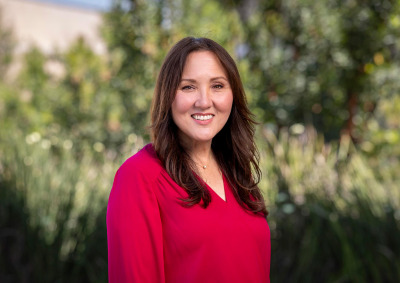A professional headshot of a smiling woman with long brown hair, wearing a vibrant magenta V-neck blouse. She is positioned against a soft-focus background of lush green foliage and trees.