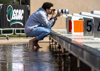 A barefoot photographer in a grey hoodie and jeans crouches on a wet pool deck, aiming a Canon camera with a large telephoto lens toward the starting blocks. In the background, a banner reads "GSAC."