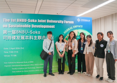 A man stands next to six women, all wearing nametags, as they pose in front of a large green and blue sign reading "The 1st BNBU-Soka Joint University Forum on Sustainable Development"
