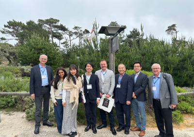 A group of conference attendees wearing formal attire pose outside in front of various greenery on Asilomar Conference Grounds in Pacific Grove, California.
