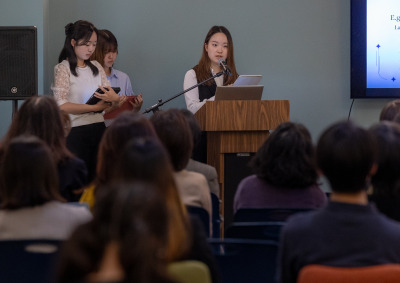 Three people wearing professional attire stand at the front of the room. One is speaking into a microphone at a wooden lectern to a seated audience.