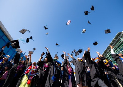 SUA Graduates wearing a mix of colorful clothes under their black graduation gowns throw their caps into the air in celebration.