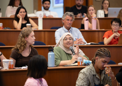 A woman wearing a tan headscarf and green shirt speaks into a microphone as people sitting around her listen on.