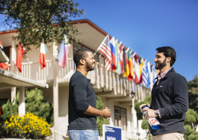 Two men wearing black longsleeve shirts smile as they converse outside a two-story building with various countries' flags hanging over the balcony.