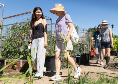 Four people wearing casual attire walk on a dirt path through rows of gardening plots with a lot of greenery.