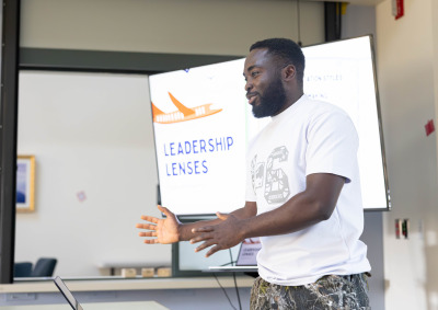 A man wearing a white t-shirt and camo pants gestures with his hands as he stands in front of a projector screen that reads, "Leadership Lenses."