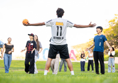 A student with short brown hair wearing a white Neymar jersey stands with his back to the camera and arms outstretched, holding a dodgeball in one hand as other dodgeball players look on.