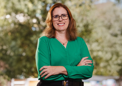 A woman with reddish-brown, shoulder-length hair is wearing a light green blouse, black pants, and black-rimmed glasses as she stands with her arms crossed and smiles.