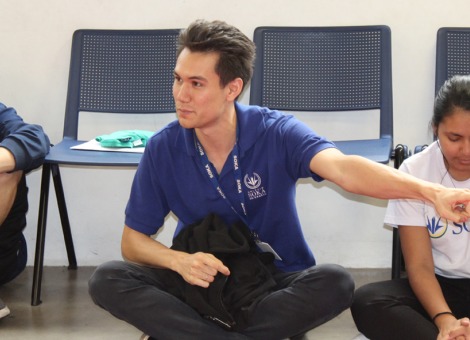 Scott Bower sits cross-legged on the floor in between two students and is pointing to his left