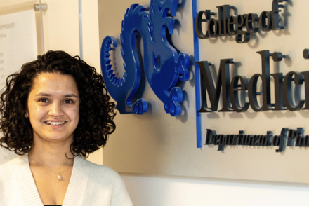Aarohee Bhattarai has short curly dark brown hair and is wearing a white shirt. She is standing to the side of wall sign that says "College of Medicine"