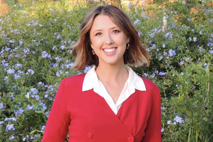 A woman with short brown hair wearing a red longsleeve shirt with a white collar poses in front of a field of blooming purple flowers.