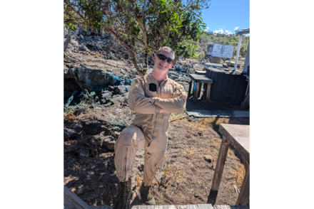 A man wearing a tan jumpsuit and sunglasses stands with his arms crossed and one boot up on a step as he works in an outdoor area.