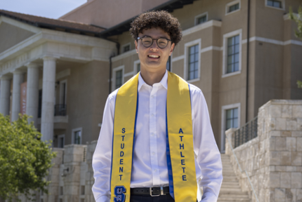 A man with curly brown hair is wearing a white dress shirt and yellow graduation stole that says "Student Athlete" as he stands in Front of a large building with white pillars.