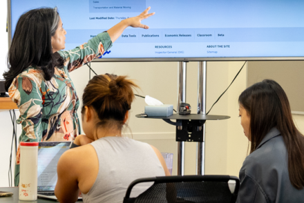 A female professor with brown hair points at a tv screen as two female students look on.