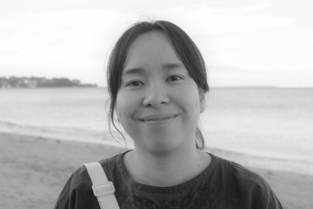 A woman with dark hair tied back in a ponytail stands in front of a body of water in a black and white photo.