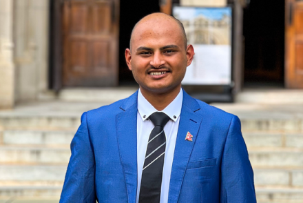 Mukesh Bastola wears a blue suit jacket and striped black tie as he stands in front of marble stairs outside a building with wooden doors.