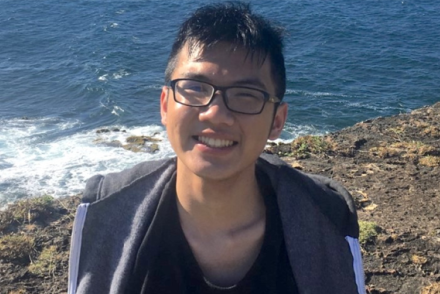 A man with short black hair and black eyeglasses smiles while standing on a cliffside overlooking the ocean