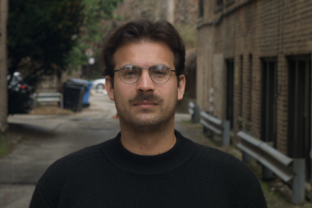 A man with short brown hair and facial hair wearing round glasses and a black longsleeve shirt poses in an alleyway lined with brick buildings.