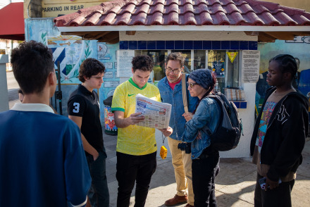 A small group of people gathers outdoors in front of a building with a red-tiled roof, looking at a printed newspaper or map held by a young man in a yellow jersey.
