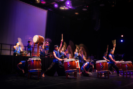Student taiko drummers in blue and black attire perform on a dark stage with purple lighting. Motion blur captures their energetic strikes on the large drums.